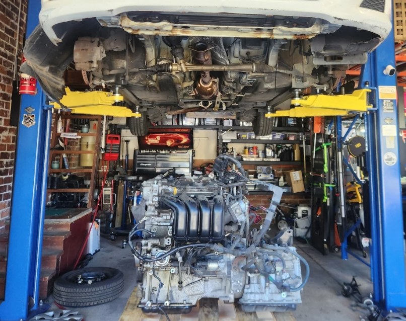 A car up on the hoist with its old engine sitting on the workshop floor